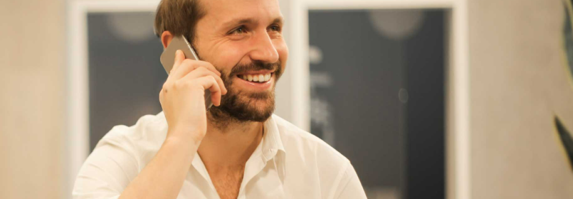 Man in witte blouse glimlacht terwijl hij aan de telefoon is in een kantooromgeving.