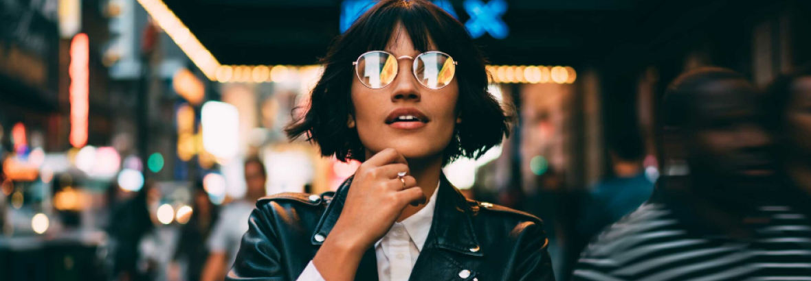 Vrouw in leren jack met reflecterende zonnebril loopt door een drukke stad vol neonlicht en voorbijgangers.